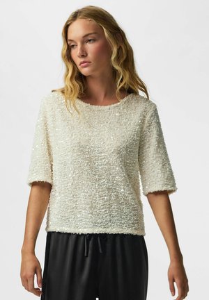 Femme aux cheveux blonds ondulés portant un haut blanc texturé à manches courtes et un pantalon noir à cordon, regardant légèrement sur le côté.