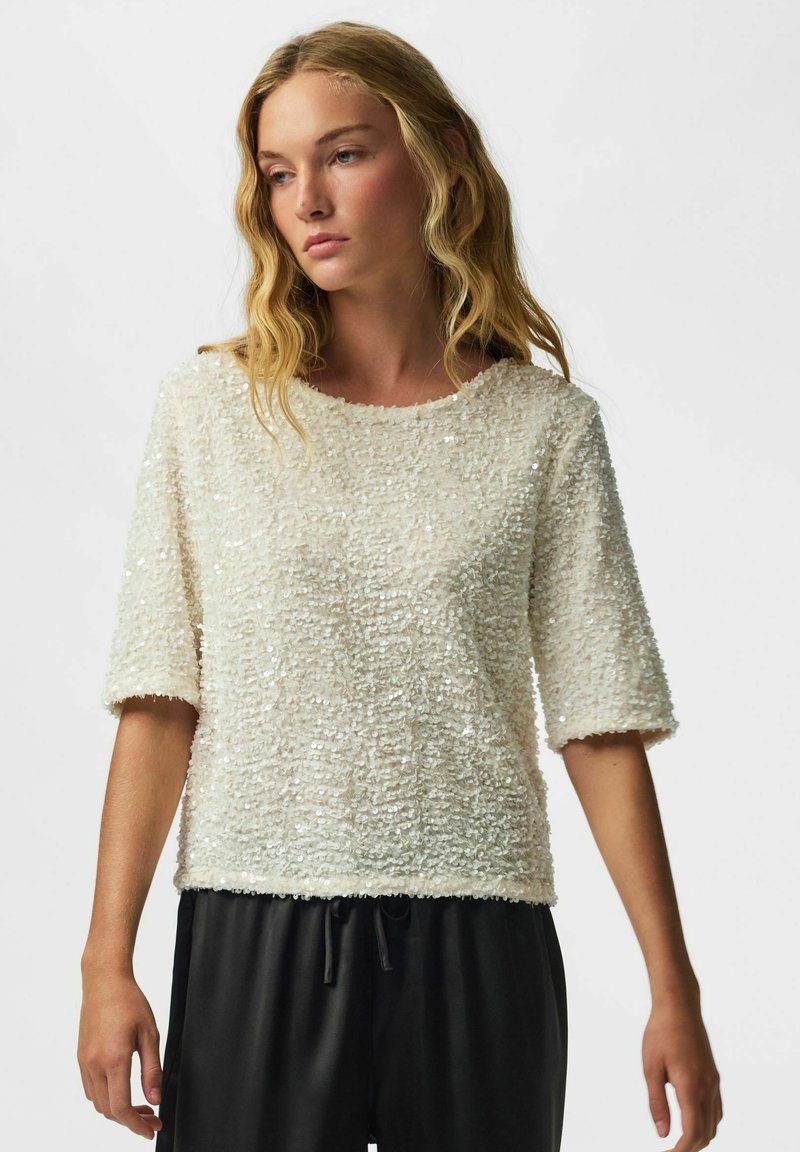 Femme aux cheveux blonds ondulés portant un haut blanc texturé à manches courtes et un pantalon noir à cordon, regardant légèrement sur le côté.