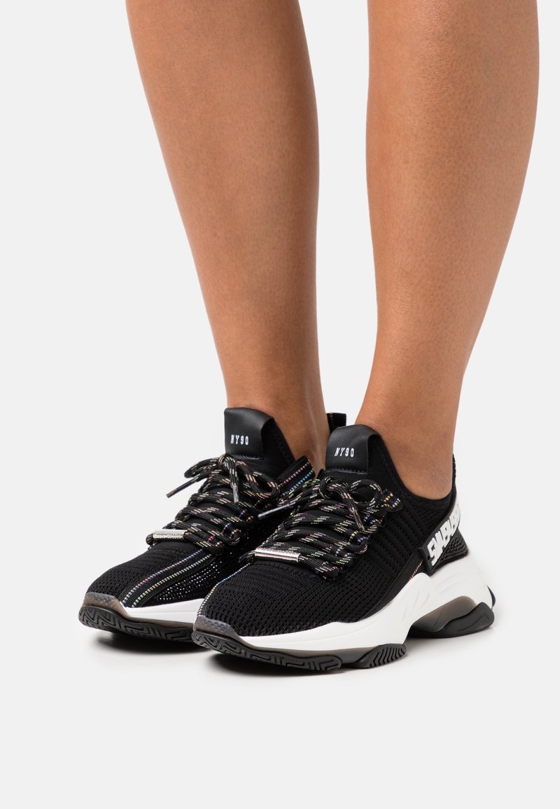 Jambes portant des baskets en maille noire avec des semelles blanches épaisses, des lacets à motif et le marquage "NY90" sur la languette et le côté.