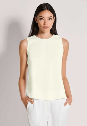 Mouwloos topje in lichtcrème, gladde stof, ronde halslijn, subtiele glans, gecombineerd met witte broek, handen in de zakken.