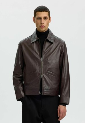 Homme portant une veste en cuir marron foncé sur un col roulé noir, debout avec une main dans la poche devant un fond clair uni.