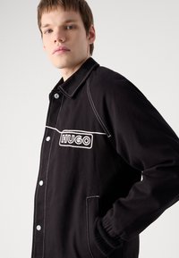 Giacca nera in tela con cuciture bianche. Presenta un logo prominente "HUGO" e tasche laterali. Design aderente e chiusure con bottoni.