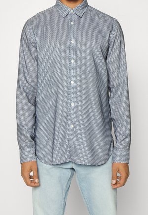 Mand iført skjorte med lange ærmer med små marineblå geometriske mønstre og lyseblå jeans.