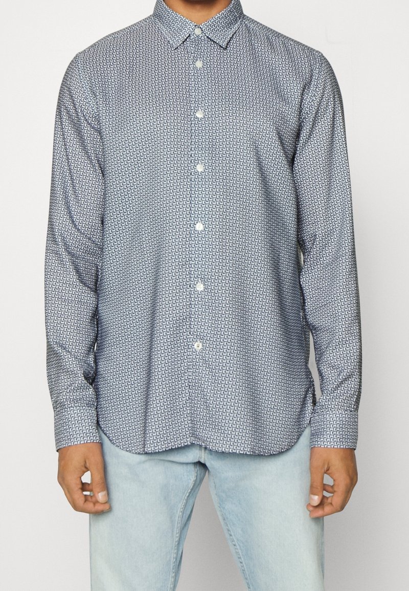 Homme portant une chemise à manches longues boutonnée avec un petit motif géométrique bleu marine et un jean bleu clair.