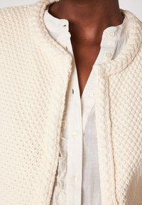 Close-up van een persoon die een textuurrijke crèmekleurige cardigan draagt over een witte blouse met knopen, met gerafelde details bij de kraag en aan de voorkant.