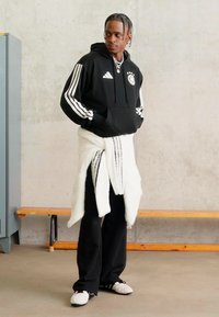 Mężczyzna w czarnej bluzie Adidas, czarnych spodniach, białym swetrze przewiązanym w talii i białych trampkach, stojący w pomieszczeniu obok drewniej ławki i szafki.