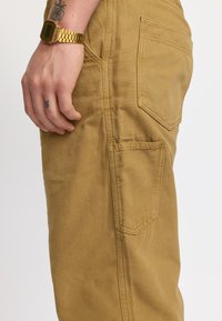 Pantaloni utility color khaki con tasche laterali rinforzate, tessuto di cotone testurizzato e vista laterale che mostra un orologio digitale dorato al polso.