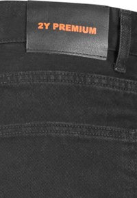 Jeans de mezclilla negra con una etiqueta de cuero en la cintura que lleva el texto "2Y PREMIUM" en relieve en color naranja. La tela tiene una textura suave y un brillo sutil.
