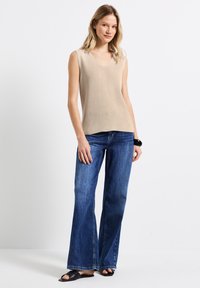Top beige senza maniche con scollatura a V, texture a coste, abbinato a jeans a zampa blu e sandali neri a scivolo. Il modello è in piedi davanti a uno sfondo semplice.
