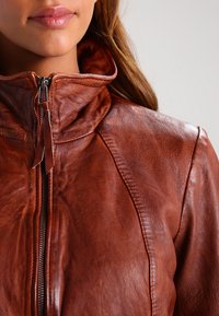 Brun läderjacka med hög krage, med en framdragkedja, mjuk textur och synliga sömdetaljer längs sömmarna.