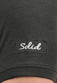 Mørk grøn stof t-shirt tæt på med en struktureret overflade. En hvid etiket syet med ordet "Solid" er synlig.