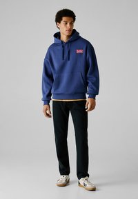 Homem jovem vestindo um hoodie azul com o logótipo da Red Bull Racing, calças pretas e ténis brancos, em pé contra um fundo cinzento liso.