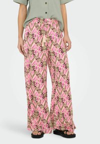 Femme portant un pantalon à jambes larges avec un motif abstrait rose, vert et marron, taille à cordon, associé à un chemisier vert à manches courtes et des sandales noires.
