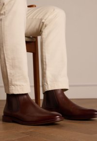 Bottes Chelsea en cuir marron avec des panneaux élastiques sur les côtés, texture lisse et bout arrondi, associées à un pantalon blanc, assises sur une chaise en bois.