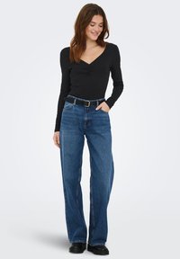 Sort langærmet top med V-udskæring og gathered detalje, kombineret med vide blå jeans i denim og sort bælte. Glat tekstur, afslappet pasform.