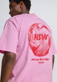 Lyserød t-shirt med et stort rødt grafisk print af en fodbold med teksten "serve the game" og "hummel sportswear" nedenunder.