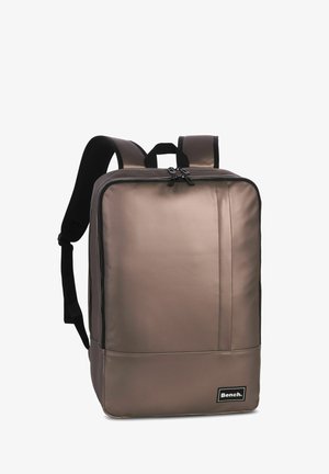 Bruine rugzak met een gladde, matte textuur, voorzien van twee zwarte verstelbare banden, een handvat aan de bovenkant en een logo-patch aan de voorkant.