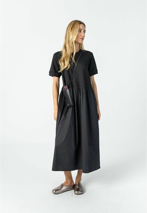 Kvinde med langt blondt hår iført en sort midi-kjole med korte ærmer, metalliske slip-on sko og en sort crossbody telefonpung.