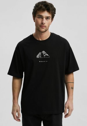 Mężczyzna z kręconymi włosami, ubrany w czarną oversize koszulkę z białym geometrycznym wzorem budynku i napisem „GDYNIA”, stoi na jednolitym tle.