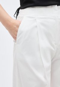 Χέρι που έχει τοποθετηθεί στην τσέπη άσπρου κομμένου παντελονιού, που φοριέται με μαύρο τοπ, εμφανίζοντας την υφή του υφάσματος και τις λεπτομέρειες της πτυχής.