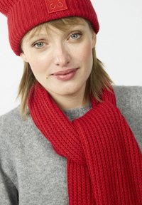 Bonnet en tricot rouge avec un petit logo, associé à une écharpe en côtes rouges assortie, porté sur un pull gris clair.