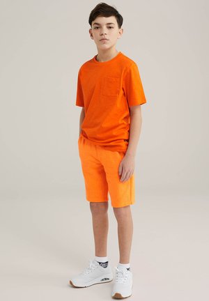 Ljus orange kortärmad T-shirt med bröstficka och matchande orange shorts. Vita sneakers med strukturerade sålar fulländar outfiten.