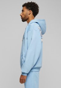 Light blue hoodie made of soft fabric, featuring a large hood, kangaroo pocket, and graphic design on the side. Standard fit with ribbed cuffs.