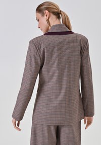 Bruin en beige houndstooth blazer met een notch-kraag, lange mouwen en een effen bordeauxrode accent bij de kraag. Zichtbare naaddetails.