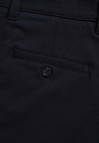 Gros plan d'une poche arrière en tissu noir d'un pantalon avec un seul bouton noir et une passants de ceinture à la taille.