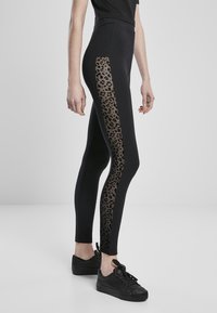 Svarte leggings med en leopardtrykt, gjennomsiktig panel på ytre lår. Har høy midje og tettsittende design. Brukes med svarte sneakers.