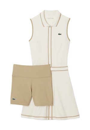Vestito bianco senza maniche con dettagli beige e una cerniera, abbinato a pantaloncini beige. Presenta un logo Lacoste verde su entrambi i capi.