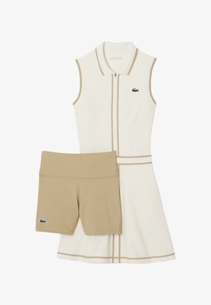 Vestito bianco senza maniche con dettagli beige e una cerniera, abbinato a pantaloncini beige. Presenta un logo Lacoste verde su entrambi i capi.
