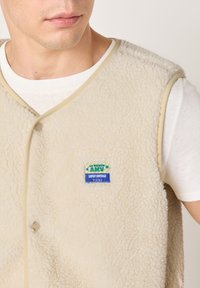 Smėlio spalvos fliso liemenė, turinti minkštą tekstūrą, apvalų iškirptę, priekinį sagų užsegimą ir spalvingą prekės ženklo lipduką ant krūtinės.