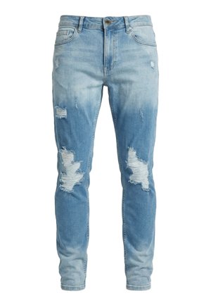 Homme portant un jean bleu clair déchiré et des baskets blanches, debout jambes croisées sur fond blanc.