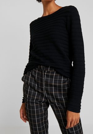 Personne portant un haut noir à manches longues côtelé et un pantalon noir avec des motifs en grille blanche et brune, debout devant un fond clair uni.