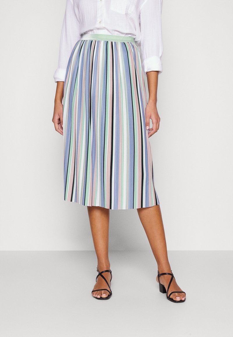 Femme portant une jupe midi plissée à rayures verticales pastel, associée à une chemise blanche à manches longues et des sandales noires à brides.