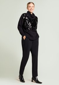 Suéter negro de punto con cuello bote y adornos de lentejuelas en la parte delantera. Combinado con pantalones negros a medida y botines de tacón.