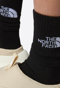 Calcetines negros acanalados con el logo "THE NORTH FACE" en blanco, combinados con calzado deslizable beige con un upper texturizado y una pestaña en el talón.
