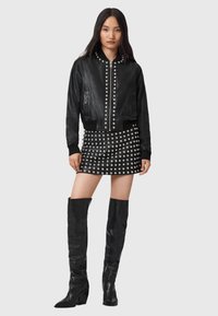 Giubbotto bomber in pelle nera con dettagli in borchie argentate, abbinato a una mini gonna nera con accenti di borchie e stivali neri alti fino al ginocchio.