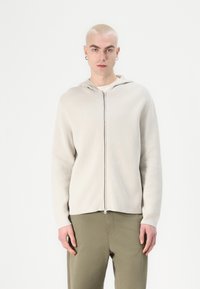 Ljusbeige zip-up hoodie i mjukt tyg med huva, med framzipper och ribbade muddar. Bärs över en ljus topp och gröna byxor.
