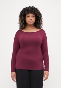 Burgundy long-sleeve top with a wide scoop neckline, made of soft fabric, featuring a fitted silhouette and smooth texture. Worn with black pants.