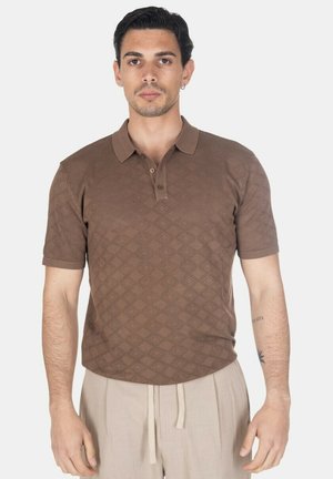 Uomo in piedi rivolto in avanti che indossa una polo marrone a maniche corte con motivo e pantaloni beige con coulisse su uno sfondo neutro.