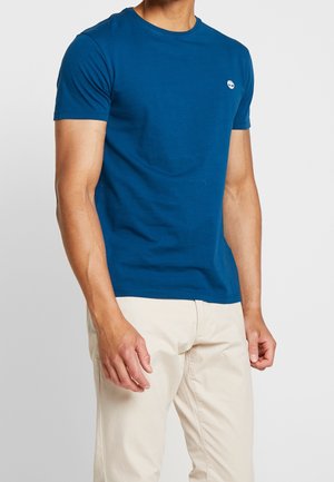 T-shirt en coton bleu avec un col rond et des manches courtes. Présente un petit logo sur la poitrine. Portée avec un pantalon de couleur claire.