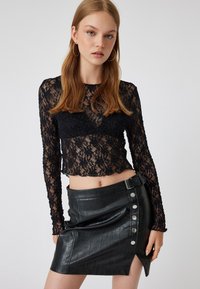 Schwarzes Spitzen-Crop-Top mit langen Ärmeln und floralem Muster, kombiniert mit einem hoch taillierten, schwarzen Minirock aus Kunstleder mit silbernen Knopfakzenten.