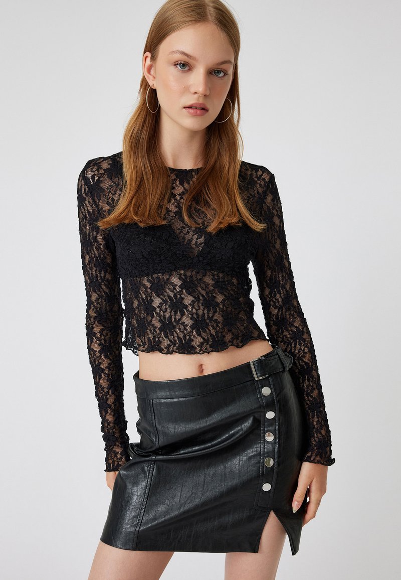 Schwarzes Spitzen-Crop-Top mit langen Ärmeln und floralem Muster, kombiniert mit einem hoch taillierten, schwarzen Minirock aus Kunstleder mit silbernen Knopfakzenten.