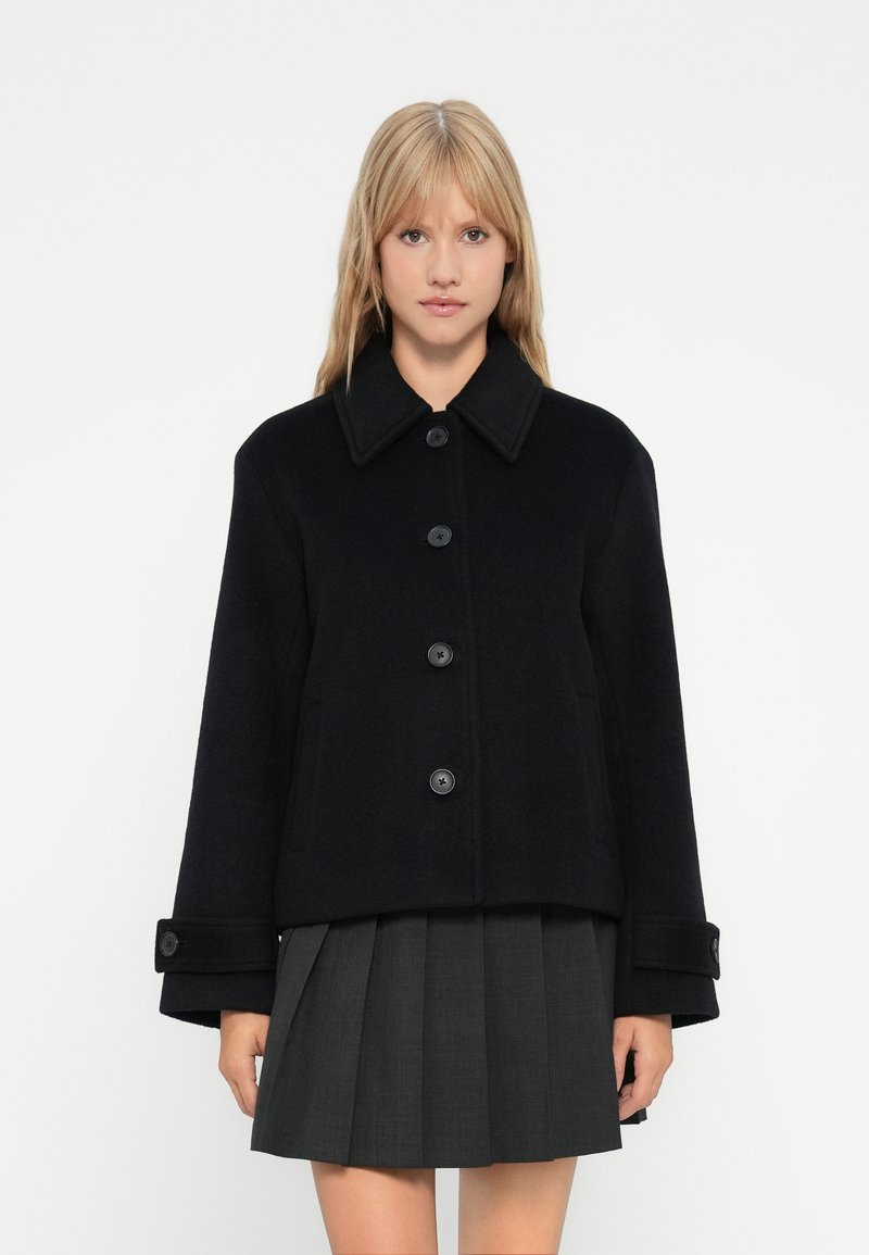 Žena s plavom kosom koja nosi crnu jaknu na dugmad i tamnosivu suknju s nabranicama, stoji ispred obične bijele pozadine.