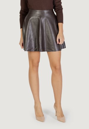 Donna che indossa una gonna in pelle marrone scuro, una maglietta a maniche lunghe marrone scuro e scarpe con tacco nude, in piedi con le braccia rilassate.