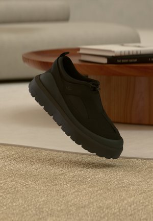 Baskets noires épaisses à semelle épaisse flottant au-dessus d'un tapis beige, avec une table basse en bois et des livres empilés dans un salon.