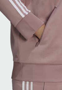 Chaqueta deportiva rosa con rayas blancas, puños acanalados y detalle de bolsillo con cremallera. Cuenta con tela suave y logotipo de marca.
