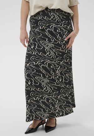 Moteris dėvi juodai baltos abstraktaus rašto ilga sijoną su juodais smailiais kulniukais ir šviesiai smėlio spalvos viršų.